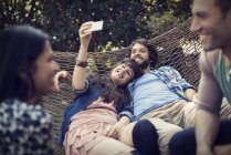 Group of cheerful friends lounging in hammock in garden, talking and taking selfie. — Stock Photo