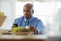 Mann im blauen Hemd sitzt mit Kaffee am Frühstückstisch und benutzt Smartphone. — Stockfoto