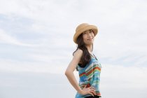Mujer asiática alegre con sombrero de paja de pie al aire libre contra el cielo nublado
. - foto de stock