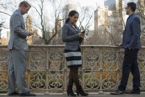 Tres personas de negocios revisando teléfonos inteligentes mientras están de pie en la balaustrada en el parque de la ciudad . - foto de stock