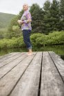 Низкий угол обзора женщины, стоящей на причале у озера и держащей чашку кофе . — стоковое фото