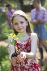 Niña de edad elemental sosteniendo planta con follaje verde en las manos con hermanas en el fondo . - foto de stock
