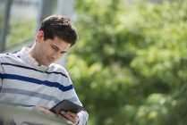 Молодий чоловік спирається на перила в парку і використовує цифровий планшет . — стокове фото