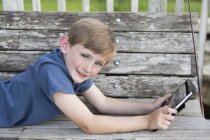 Elementary age boy lying on front at wooden bench outdoors with digital tablet. — Stock Photo