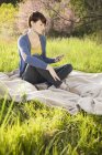 Молода жінка сидить у трав'янистому полі на ковдрі і використовує цифровий планшет . — стокове фото