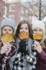 Trois adolescentes tenant des feuilles automnales devant des visages . — Photo de stock