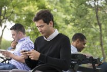 Група людей, які сидять в парку і використовують смартфони на лавці . — стокове фото