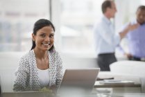 Young woman smiling while sitting at desk with laptop computer with men talking in background. — Stock Photo