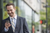 Бизнесмен в костюме стоит на улице, улыбается и пользуется смартфоном . — стоковое фото