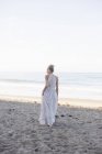 Blonde woman in long dress standing on sandy beach. — Stock Photo