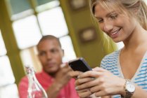 Frau checkt Smartphone mit Mann im Hintergrund in Café. — Stockfoto