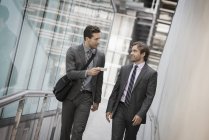 Dos hombres de negocios en trajes fuera del edificio sosteniendo teléfono inteligente y hablando . - foto de stock