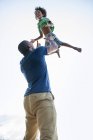 Vista de ángulo bajo del padre jugando y levantando niños de edad elemental al aire libre . - foto de stock