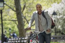 Mid adult man pushing bicycle in sunny park. — Stock Photo