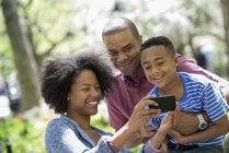 Dois pais e menino tirando selfie com smartphone no parque ensolarado . — Fotografia de Stock