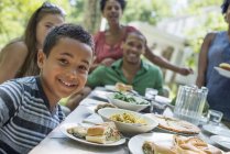 Niño sonriendo en la mesa con la familia reuniéndose alrededor de la mesa de la cena en el jardín rural . - foto de stock