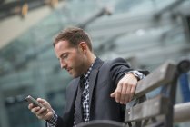 Мужчина в деловом костюме с короткими волосами с помощью смартфона на уличной скамейке . — стоковое фото