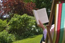 Человек, сидящий в шезлонге и читающий книгу о зеленой лужайке . — стоковое фото