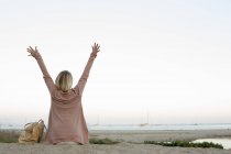 Blonde woman sitting on sandy beach with arms raised. — Stock Photo