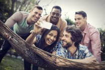 Group of cheerful friends lounging in hammock in garden and taking selfie. — Stock Photo