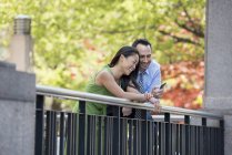 Man and woman checking smartphone while leaning on fence under trees in park. — Stock Photo