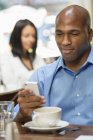 Mid adult man using smartphone in coffee shop with woman in background. — Stock Photo