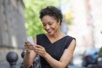 Mujer en vestido negro revisando el teléfono en la calle de la ciudad . - foto de stock
