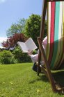 Pessoa sentada em cadeira de praia e livro de leitura no gramado verde . — Fotografia de Stock
