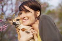 Young woman holding and hugging chihuahua dog in park with spring blossom. — Stock Photo