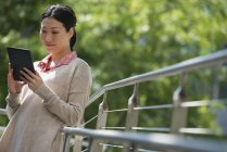 Asian woman leaning on railing in park and using digital tablet. — Stock Photo