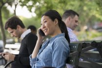 Женщина по телефону сидит на скамейке в парке с людьми, использующими телефоны . — стоковое фото