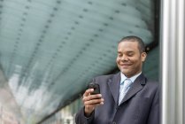 Empresário de terno em pé na rua, sorrindo e usando smartphone . — Fotografia de Stock