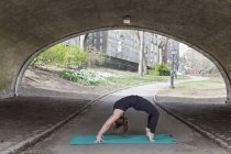 Donna inarcando di nuovo sotto il ponte sul tappetino yoga a Central Park . — Foto stock