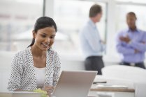 Young woman sitting at desk and using laptop in office with coworkers in background. — Stock Photo