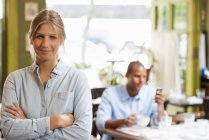 Woman standing with arms folded in cafe interior with man using phone in background. — Stock Photo