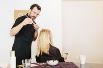 Мужчина-парикмахер с использованием фольги при окрашивании светлых волос клиента-женщины со смартфоном . — стоковое фото