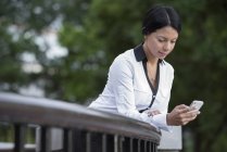 Frau in weißer Jacke lehnt an Holzgeländer und checkt Smartphone. — Stockfoto