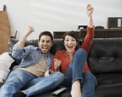 Homme et femme assis, célébrant et pompant l'air avec les poings tout en regardant la télévision sur le canapé . — Photo de stock