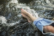 Vue recadrée de la femme en jeans à la mode avec des pieds de refroidissement déchirure dans l'eau courante de la rivière . — Photo de stock