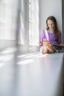 Mädchen im Grundalter sitzt auf Fensterbank und liest Buch. — Stockfoto