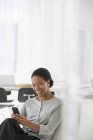 Mujer de negocios alegre utilizando el teléfono inteligente en silla cómoda y mirando en la cámara en la oficina . - foto de stock