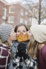 Teenage girls kissing on cheek s friend with autumn leaf in front of face. — Stock Photo