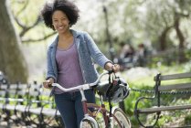 Mujer adulta mediana con peinado afro en ropa casual empujando bicicleta en el parque  . - foto de stock