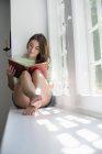 Young woman sitting on window sill and reading book. — Stock Photo