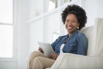 Mid adult woman sitting in armchair with digital tablet and smiling. — Stock Photo