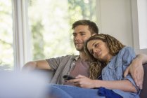 Casal com controlador remoto sentado lado a lado abraçando e assistindo tv dentro de casa . — Fotografia de Stock