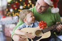 Padre e hijo sentados junto al árbol de Navidad y tocando la guitarra . - foto de stock