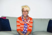 Femme de race mixte avec des cheveux blonds dans des lunettes rondes assis sur le canapé et riant . — Photo de stock