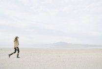 Donna dai capelli lunghi e ricci che cammina sulla spiaggia sabbiosa indossando cappello e stivali di pelle . — Foto stock