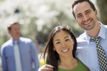 Couple standing side by side and smiling in camera with man in background. — Stock Photo